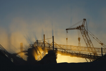 Silhouettes constructions of stone crushing equipment at a mining enterprise in a cloud of dusty illuminated by the setting sun. Quarry mining machinery.