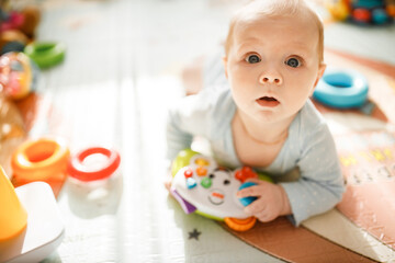 Beautiful cute toddler laying on colorful mat at home. Adorable baby girl play in kids room, looking at the camera with big interest. Childhood and childcare concept