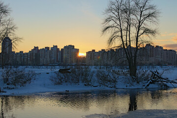 Obraz premium Picturesque winter landscape of frozen Dnieper River during sunset. Buildings of The Obolon neighborhood in the background. Bare tree reflected in the water. The sun is hiding behind the house. Kyiv