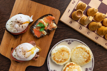 Breakfast and chess. Homemade poached eggs, sandwich with salmon fish, cheese pancakes and chess game