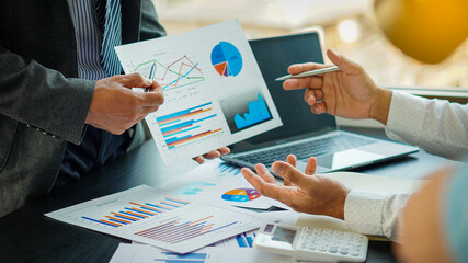 Two businessmen were analyzing a cost graph on a desk in a conference room with a calculator and a laptop with orange light.