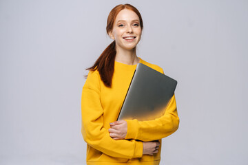Smiling young woman student holding laptop computer and looking at camera on isolated gray...