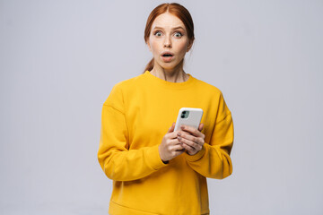 Portrait of shocked cute young woman holding mobile phone and looking at camera on isolated white background. Pretty redhead lady model emotionally showing facial expressions in studio, copy space.