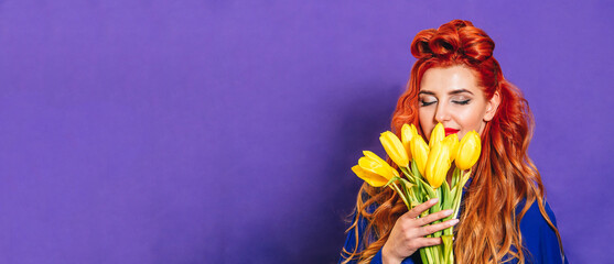 Beautiful girl with red hair stands on a purple background and holds a bouquet of yellow tulips. Concept of holiday - March 8, international women's day, Mother's Day. Close-up. Banner. Place for text