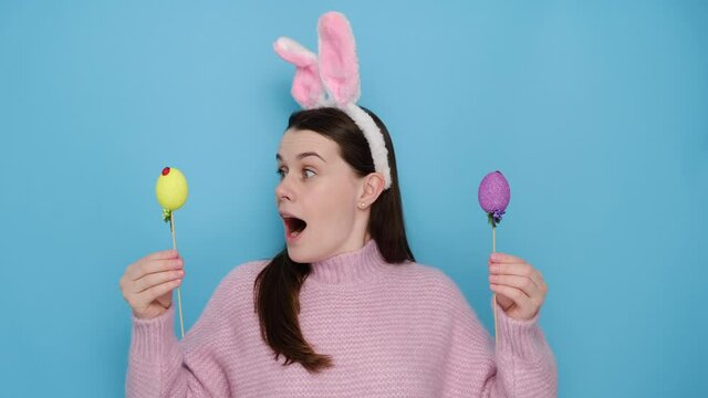 Surprised young brunette woman in bunny fluffy ears prepares for great spring festival or Easter, carries two painted eggs, joins celebration, wears pink sweater, isolated on blue studio background