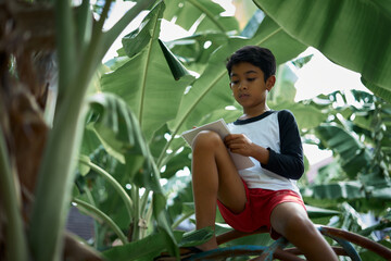 Schoolboy doing his homework while playing under banana tree in the farm 