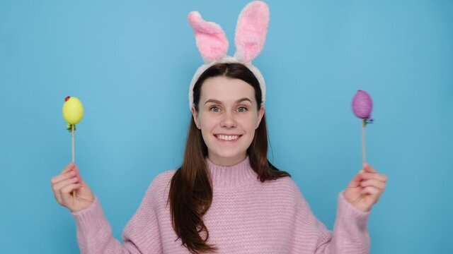 Glad caucasian young woman covers eyes with two Easter eggs, dressed in bunny ears and pink sweater, models over blue studio background with free space for promotion. Spring holiday concept