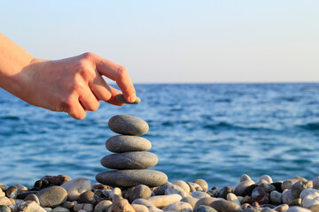 Balanced Pebbles Pyramid  on the Beach on Sunny Day and Clear Sky at Sunset. Blue Sea on Background. Woman Placing the Last Stone of Pyramid.