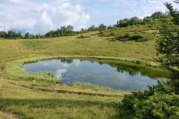 Mountain lake on the green top of the slope with the sky reflected in it with clouds.