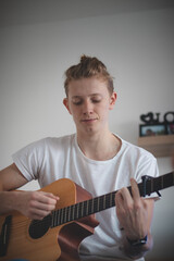 Obraz premium talented boy playing a wooden acoustic guitar in a lighted room. Blonde Man aged 20-29 strums guitar and sings from hymn book on couch. Music instruments. Music life