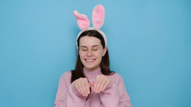 Funny young woman keeps hands as if rabbit, wears soft bunny ears, has friendly look, smiles cheerfully, ready to congratulate children with coming Easter, isolated on blue wall. I am lovely hare