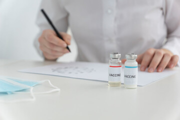 doctor's hands hold black pen and writes prescriptions for vaccination, two ampoules with vaccine stand near. Vaccination concept