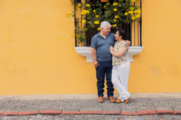 Obraz na płótnie Canvas Adult couple embracing looking at each other on a yellow wall with flowers in Antigua Guatemala- Senior couple in love on vacation in colonial city