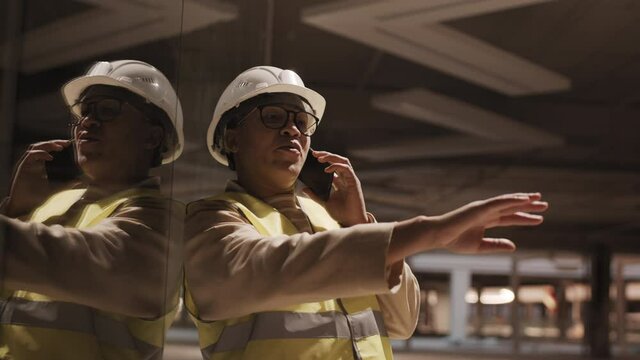 Medium close-up of female Mixed-Race construction worker wearing hard hat and glasses talking on smartphone and pointing hand forward, leaning on glass wall in dark room under renovation
