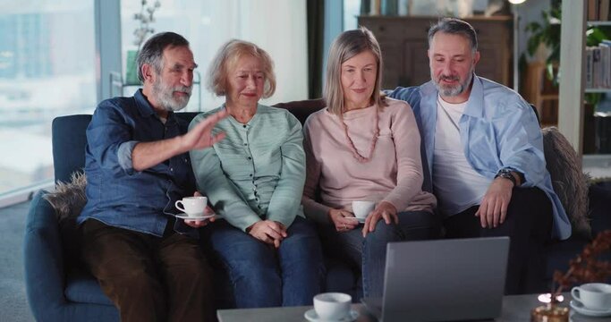 Four amazing people group of grey-haired married pensioner couples sitting on couch video conferecing on laptop computer.