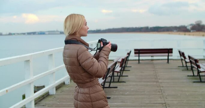 A beautiful blonde photographer takes pictures of the landscape while standing on the pier with a DSLR camera. Woman blogger travels exploring new places. Slow motion