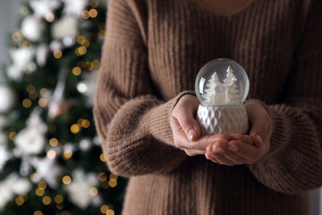Woman in sweater holding decorative snow globe against Christmas tree, closeup. Space for text