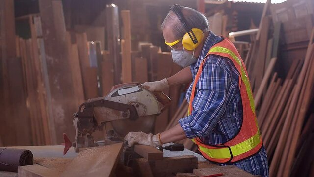 elderly carpenter craftsman in carpentry shop wearing safety personal protective equipment for protect dust, Machine noise and accident working use circular saws to cut wood board to make furniture.