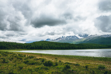 Dramatic landscape with mountain lake and forest on hills in sunlight and snowy mountains in low clouds in changeable weather. High mountains in cloudy sky and lake near green grasses and forest hills