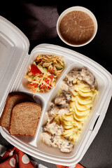 mashed potatoes with meat, salad and bread in a plastic container with coffee on a dark background top view