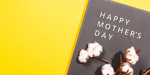 Grey plastic letter board with white quotes Happy Mother's Day, and cotton branch on a illuminating yellow background.