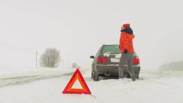 the car is parked on the side of the road on an empty road. There is a sign of attention or an emergency stop. Winter snowfall. a person dances and moves so as not to freeze. The Cold Was Crazy.