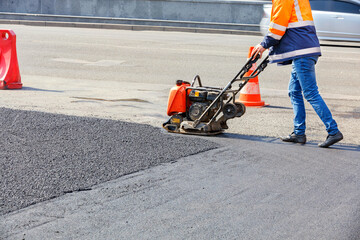 A road worker compacts asphalt with a petrol vibratory plate compactor. - obrazy, fototapety, plakaty