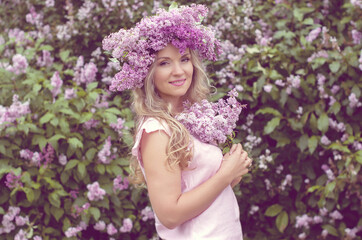 Blonde woman enjoying in a spring garden with blooming lilacs with bouquet, lilac flowers hair style. Spring Concept, Spring blossom, Positive human emotions and feelings. 