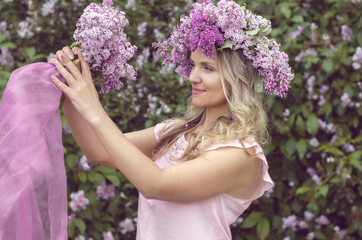 Blonde woman enjoying in a spring garden with blooming lilacs with bouquet, lilac flowers hair style.  Spring Concept, Spring blossom, Positive human emotions and feelings. 