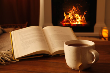 Cup of hot tea and book on wooden table near fireplace at home. Cozy atmosphere