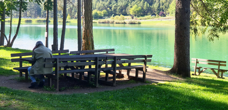 A lonely old man sits on a bench near lake in Coredo and Tavon. Panorama.
