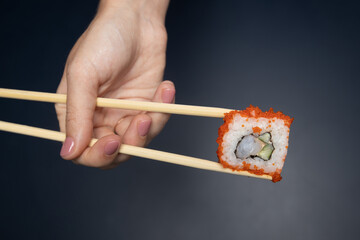 American sushi roll in female hand with chopsticks on black backdrop with space for ad. Delivery and takeaway food