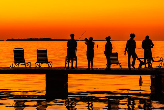 Silhouettes Of A Group Of People Fishing On A Dock At Sunset. The Sky Is Lit Bright Orange.