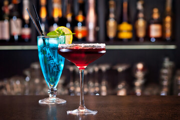 Blue and red cocktails on the bar. Alcoholic cocktails on a background of shelves with bottles. Blurred bottles of alcohol on the wall of the bar.