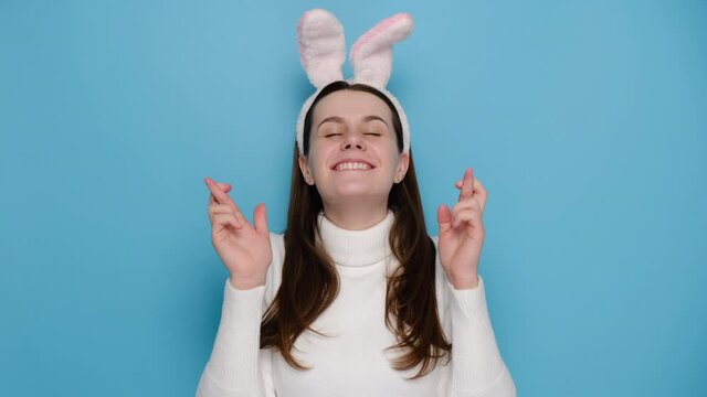 Cute young caucasian female is making wish while crossing her fingers, has positive expression, wears bunny fluffy ears and white jumper, models over blue studio background. Easter holiday concept