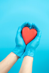 Female hands wearing blue medical gloves holding red heart over light blue background. Health care, heart disease, cardiology