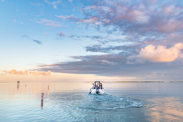 Early morning, a boat heads out on the ocean on a fishing trip in the Florida Keys as the sun rises. The ocean is calm and flat there are a few clouds in the sky. - obrazy, fototapety, plakaty