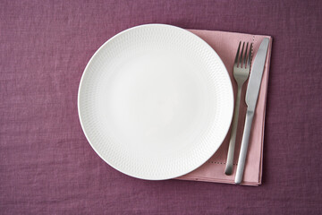 Clean empty white plate, fork and knife on bright blueberry red linen tablecloth on table, copy space, mock up, top view. Concept for menu with utensil