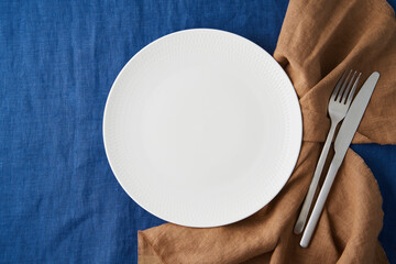 Clean empty white plate, fork and knife on bright blue dark linen tablecloth on table, copy space, mock up, top view. Concept for menu with utensil, brown textile napkin, fork and knife
