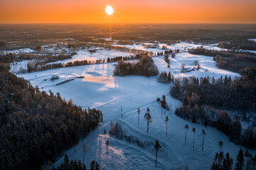 Warm sunrise over snowy countryside landscape. Pine forest covered in glowing snow. Drone aerial view.