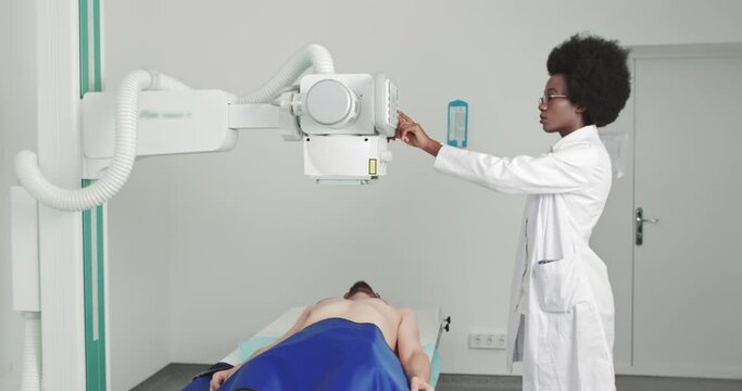 In the Hospital, Man Lying on a Bed, Female Technician adjusts X-Ray Machine. Scanning for Fractures, Broken Limbs, Injuries, Cancer or Tumor. Shot on URSA. Afro American Doctor Woman