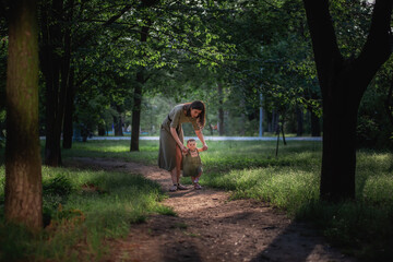 Fototapeta premium The baby girl takes the first steps, a young mother holding a child by the hands, insuring, helping to walk along the path of the park amid green trees and grass. Parental care, walks in the fresh air