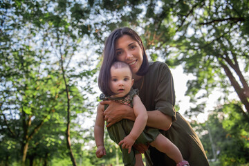 Young brown-haired mother shakes her baby in her arms in the park among the trees, green grass. Woman holds little girl in her arms, plays, hugs child who laughs. Happy childhood. Lifestyle copy space