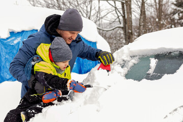 Fototapeta na wymiar Father and son removing deep snow from car. Little boy helping his father to remove snow from family car after winter attack. Father and son are removing snow and ice from their car. Copy space.