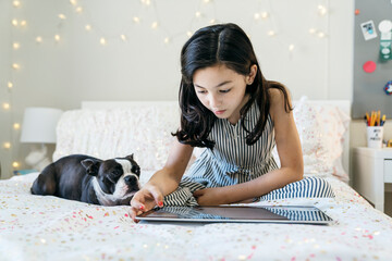 Girl sitting on bed in bedroom with Boston Terrier pet dog playing on tabled computer