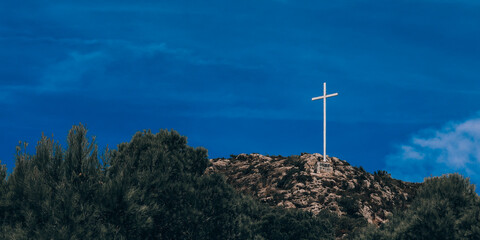 Cross on the mountain.
White cross on a mountain with trees against a blue sky, close-up side view.