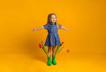 Obraz premium happy little blonde girl in a blue dress and rubber boots stands with tulips on a yellow background with space for text