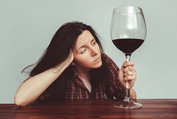A young caucasian girl in a plaid shirt with tousled hair sits at a wooden table and holds a large glass of red wine. Concept of alcohol abuse, alcoholism, hangover, loneliness and depression.