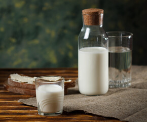 A glass of milk, a glass bottle of milk on the back, and a piece of rye bread and butter on a wooden background.