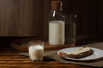 A glass of milk, a glass bottle of milk on a stand at the back, a piece of rye bread and butter on a white plate next to it.